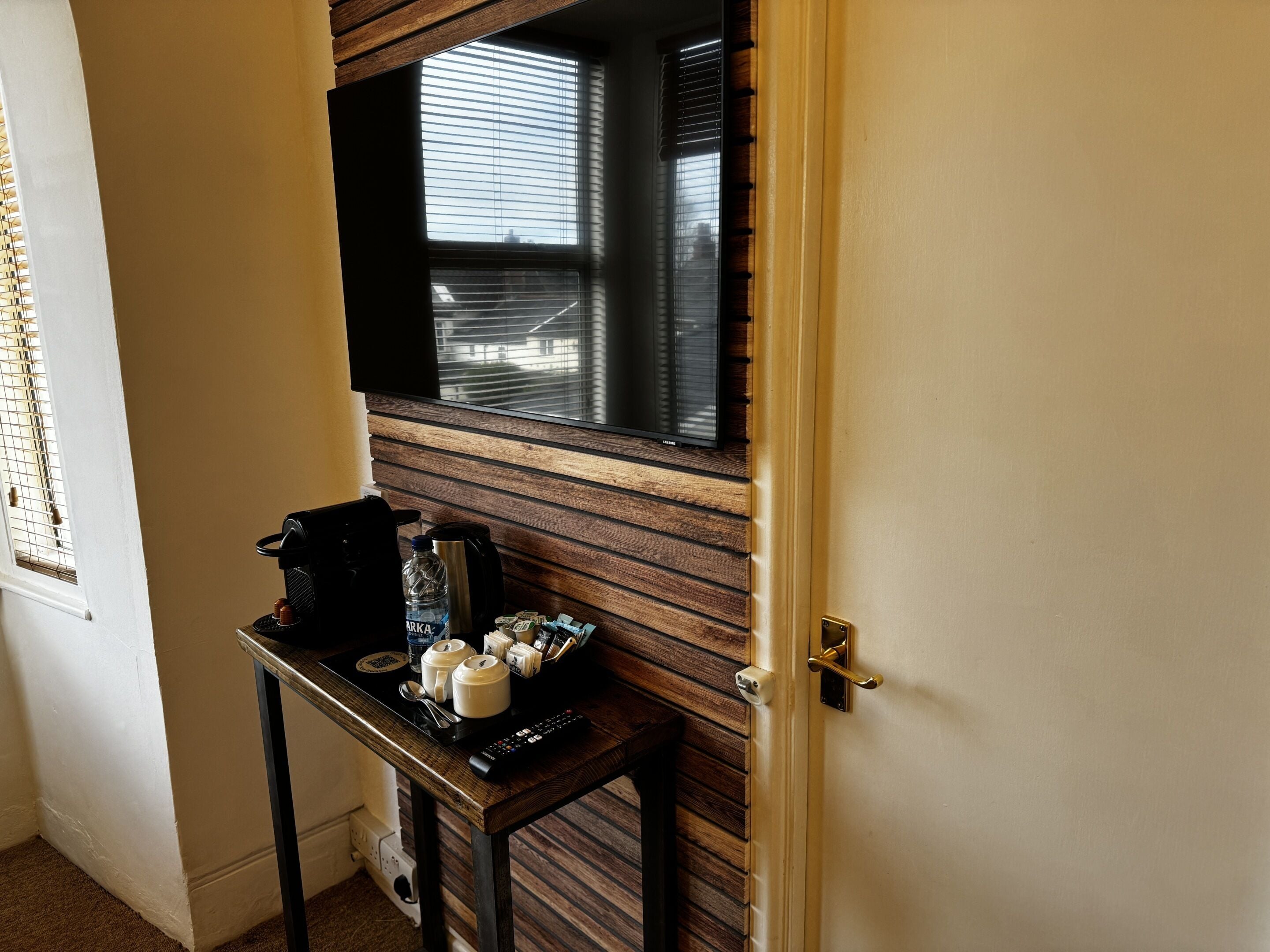 Hotelkamer met een wand van houten panelen, een klein tafeltje met een koffiezetapparaat, bekers en een afstandsbediening.