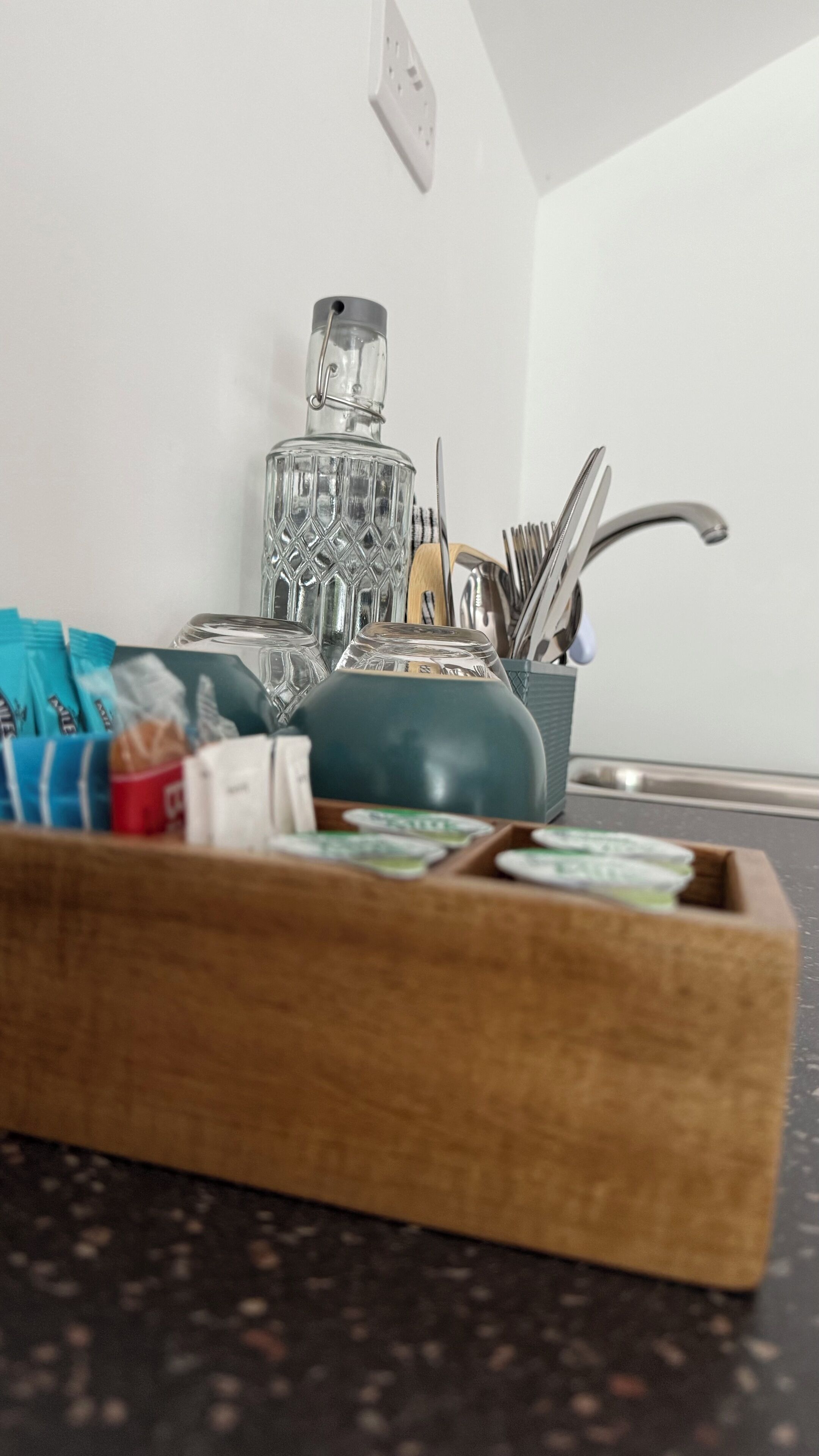 A kitchen counter with a wooden tray holding various packaged snacks, a glass bottle, and a metal container with utensils.