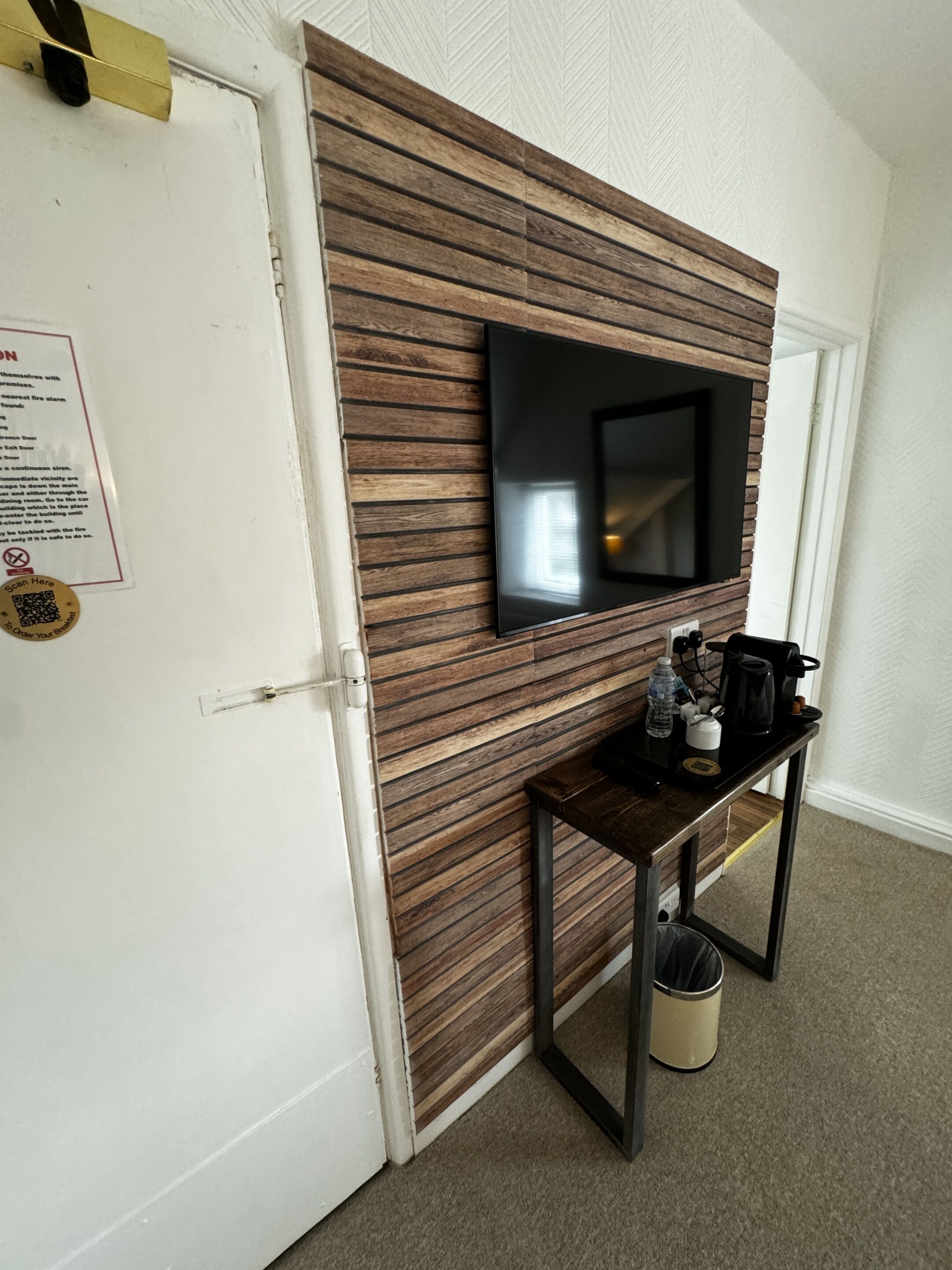 A room with a wooden panel wall, a mounted flat-screen TV, a small table with a kettle, and a trash can.