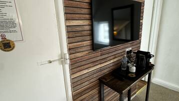 A room with a wooden panel wall, a mounted flat-screen TV, a small table with a kettle, and a trash can.
