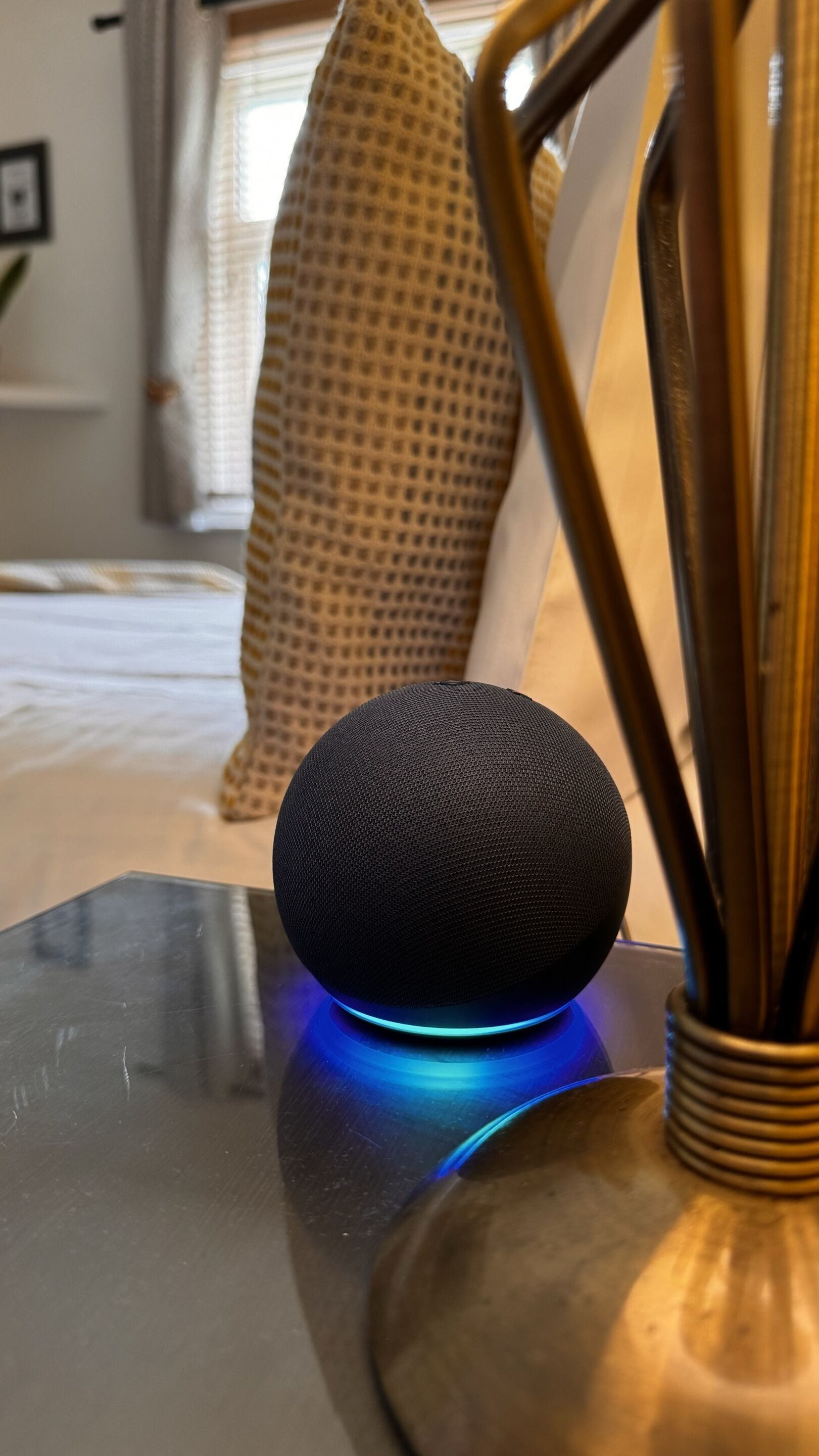 Un altavoz negro con luz azul sobre una superficie reflectante, posiblemente una mesa, en una habitación con una cama y almohadas al fondo.