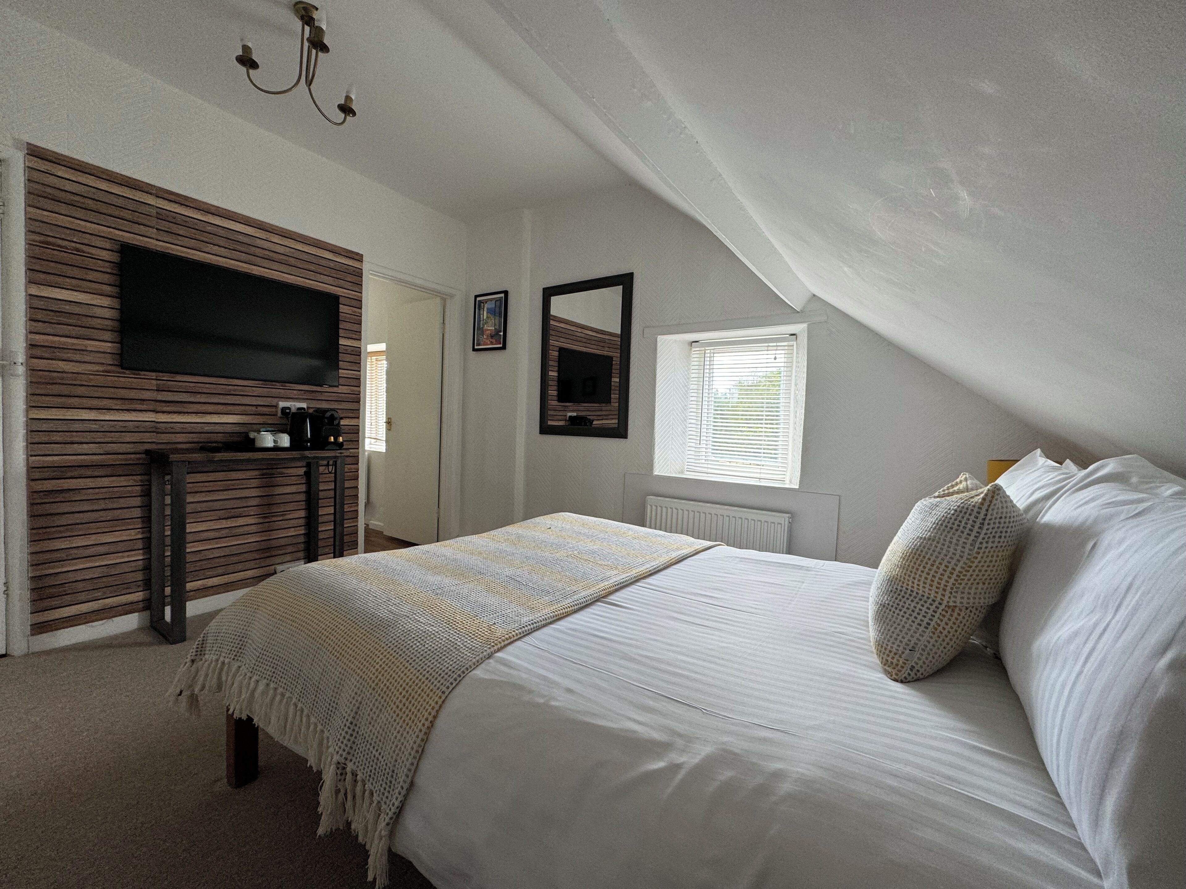 A bedroom with a bed, a television mounted on a wooden panel wall, a window with blinds, and a radiator.