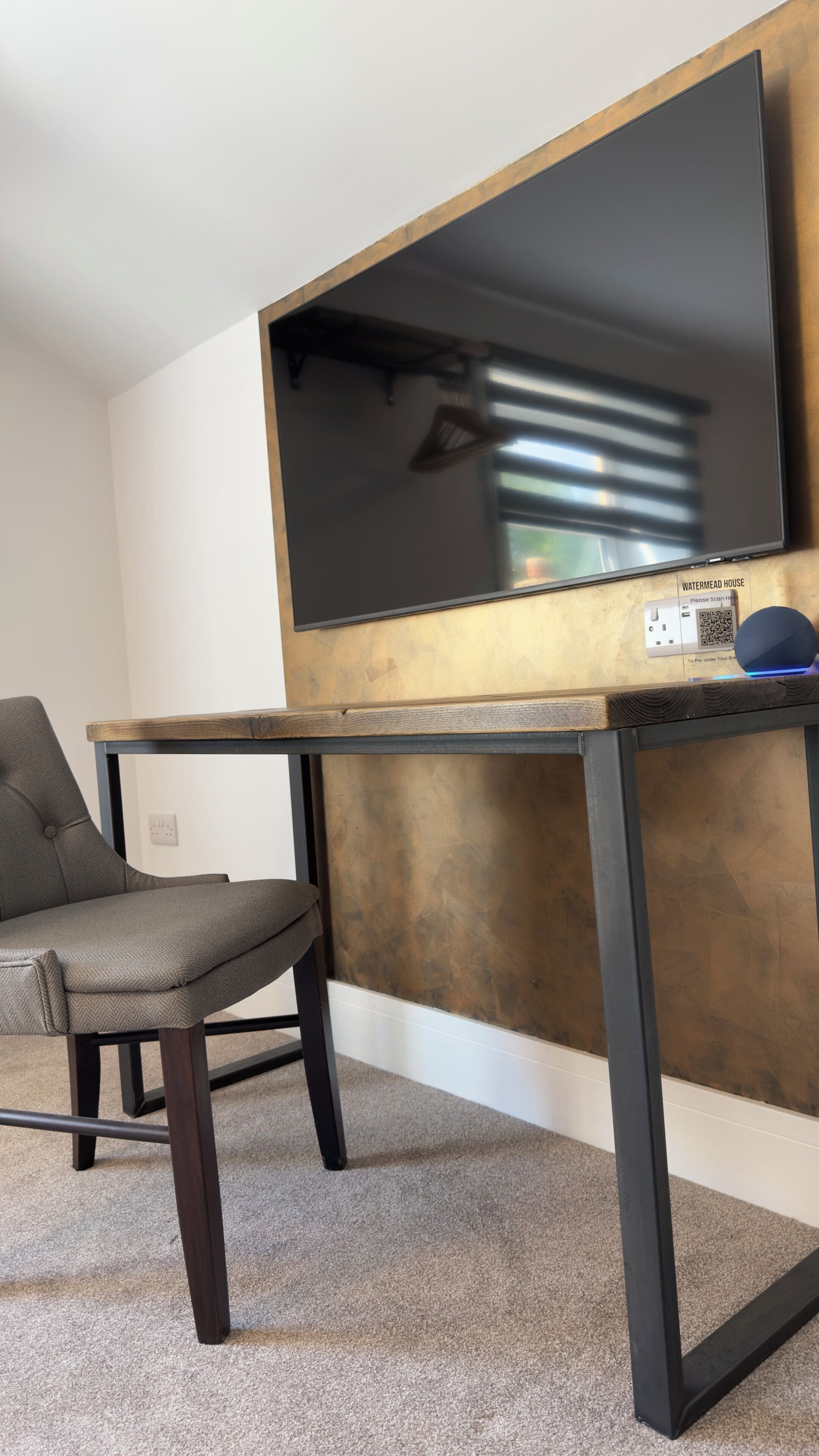Un salon moderne avec un grand téléviseur à écran plat fixé au mur, une table en bois foncé dotée d’un cadre en métal, un fauteuil recouvert d’un tissu gris et un sol recouvert d’une moquette.