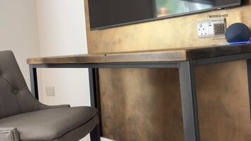 Un salon moderne avec un grand téléviseur à écran plat fixé au mur, une table en bois foncé dotée d’un cadre en métal, un fauteuil recouvert d’un tissu gris et un sol recouvert d’une moquette.