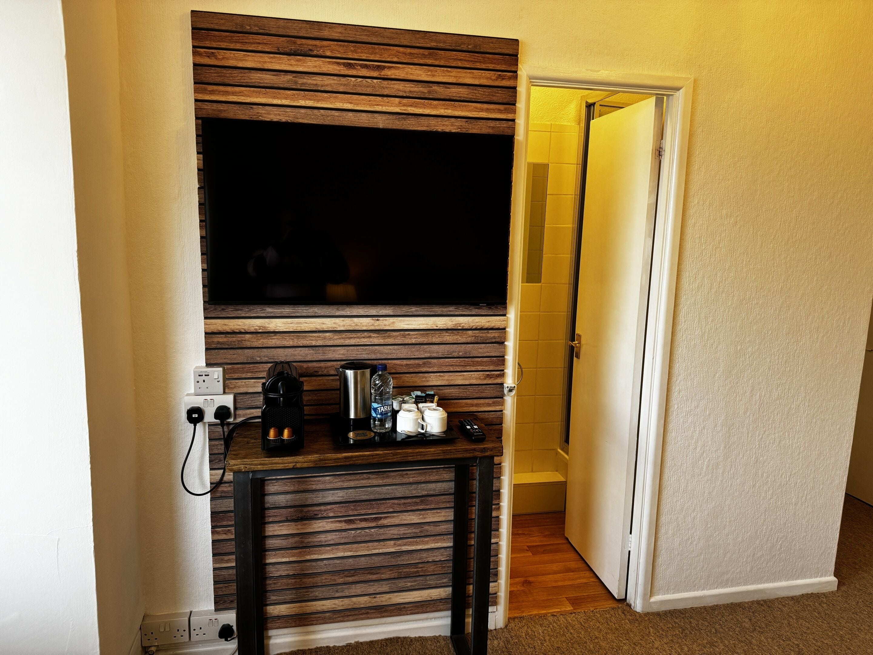 A room with a wooden slat wall, a coffee machine, a kettle, and a bottle of water on a small table.