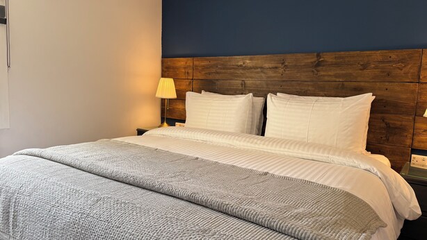 A neatly made bed with a wooden headboard, a bedside lamp, and a textured throw blanket. - Watermead House (Chard)