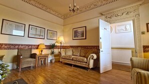 A room with a wooden floor, a patterned sofa, a chair, a lamp, and framed certificates on the wall. - Watermead House (Chard)