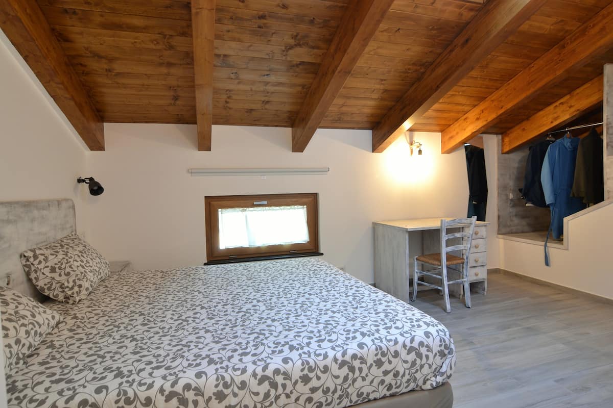 A bedroom with a bed, a desk, and a chair. There is a window and a coat rack with clothes.