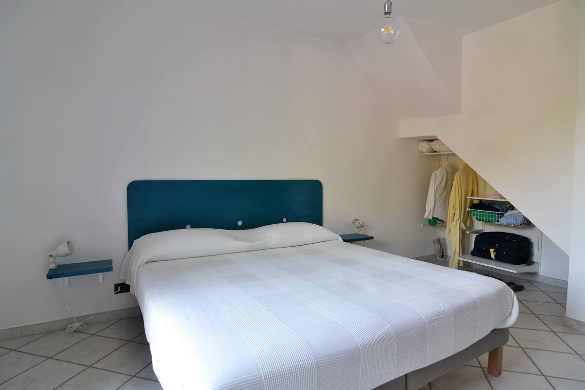 A neatly made bed with a blue headboard, a bedside table with a lamp, and a small shelf with a mirror.