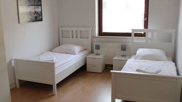 A room with two single beds, a small bedside table with a lamp, a radiator, and a window with blinds.