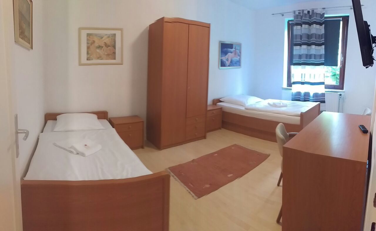 A small, tidy room with two single beds, a wooden wardrobe, a desk, and a chair.