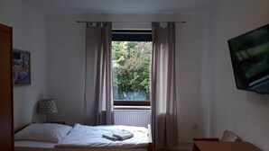 Ein Schlafzimmer mit Bett, Schreibtisch, Stuhl, Fernseher und einem Fenster mit Vorhängen.