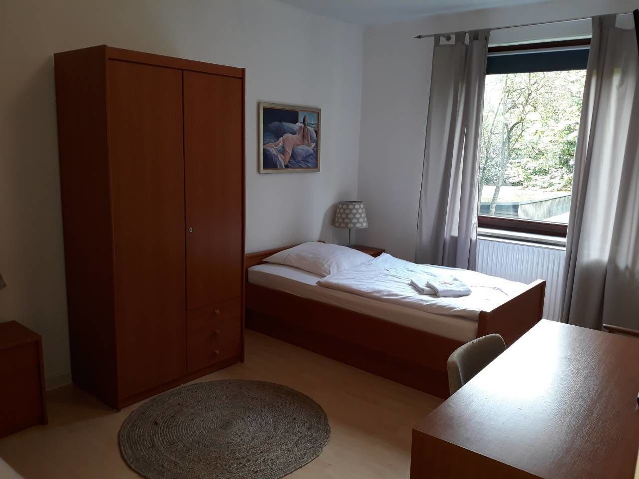 A bedroom with a bed, a wardrobe, a desk, a lamp, and a window with curtains.