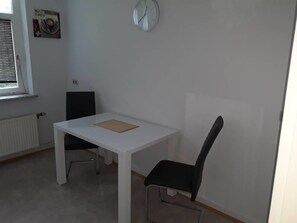 A small, sparsely furnished room with a white table and a black chair, a radiator, a window with blinds, and a clock on the wall.