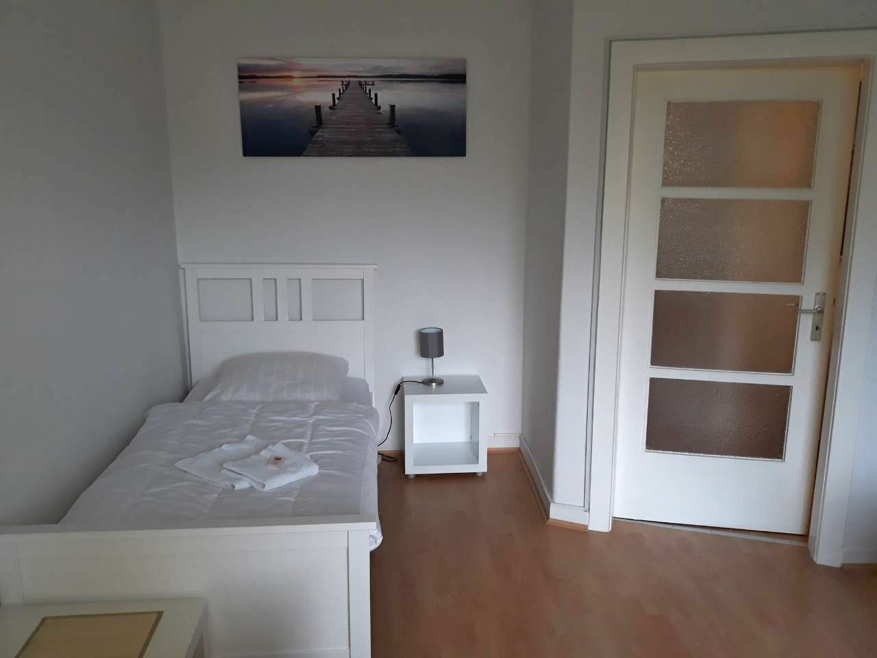 A bedroom with a bed, a nightstand, a lamp, a door, and a wall-mounted picture.