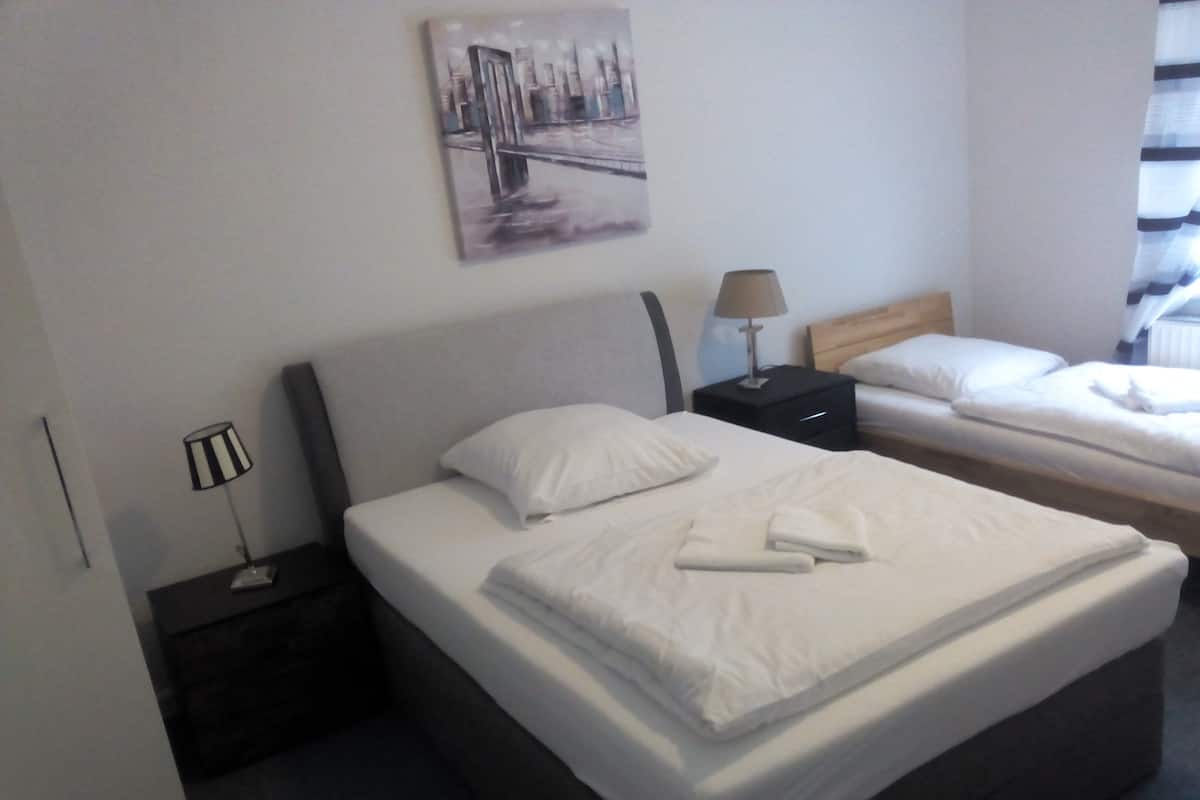 Quarto de hotel com uma cama grande, uma cama menor, uma mesinha de cabeceira com abajur e um quadro na parede.