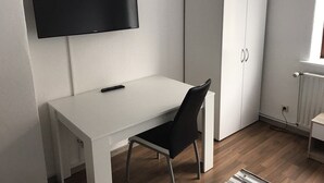 Ein schlicht eingerichteter Raum mit weißem Schreibtisch, schwarzem Stuhl, Kleiderschrank und Wand-Fernseher.