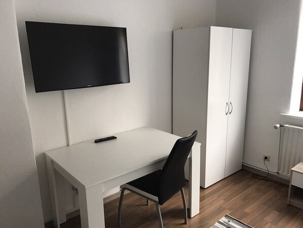 A minimalist room with a white desk, a black chair, a wardrobe, and a flat-screen TV mounted on the wall.