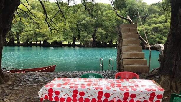 Une table recouverte d’une nappe à motifs de pommes rouges et blanches, entourée de chaises rouges, le tout se dressant sur fond d’un lac turquoise et d’arbres verdoyants.