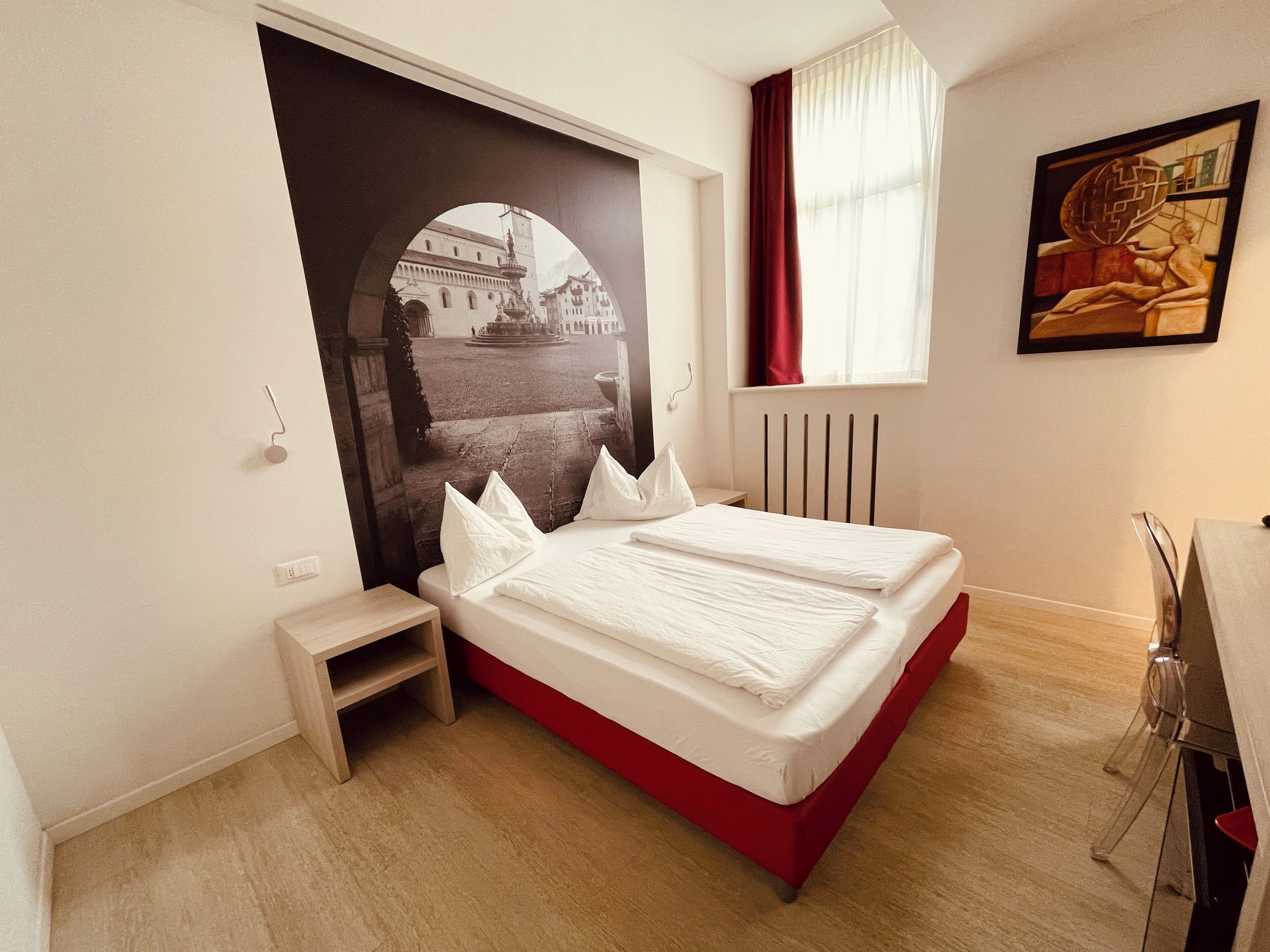A bedroom with a bed, a small bedside table, a radiator, and a window with curtains.
