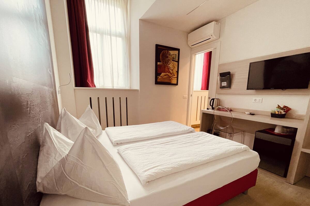 A hotel room with a bed, a desk, a chair, a TV, and an air conditioning unit.