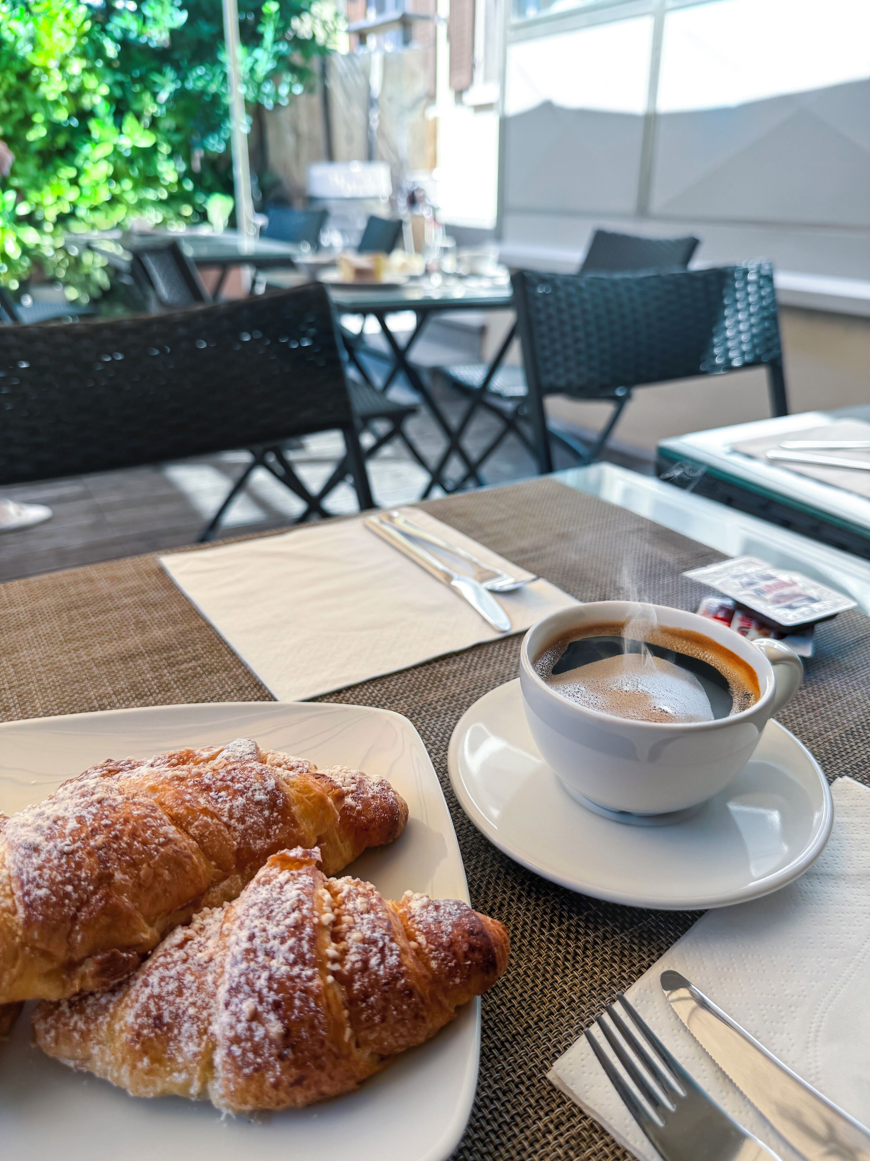 Una mesa con un plato de cruasanes y una taza de café, una servilleta y cubiertos.