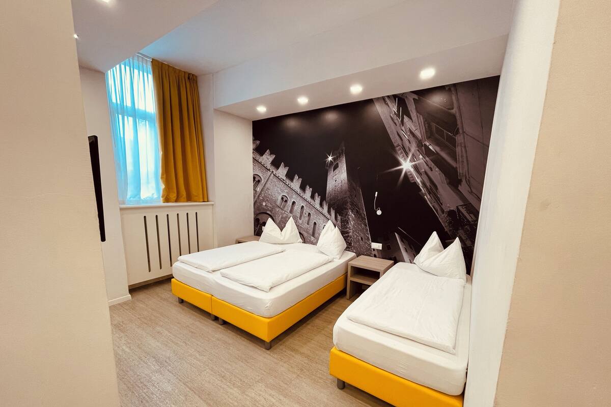 A hotel room with two beds, a window with yellow curtains, a radiator, and a wall mural of a castle.