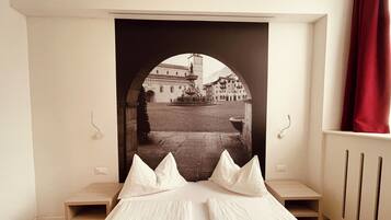 A bedroom with a bed, a small bedside table, a nightstand, and a view of a courtyard through an arched window.
