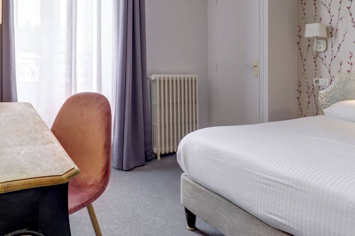 Een hotelkamer met een bed, een tafel, een stoel, een radiator en een raam met gordijnen.