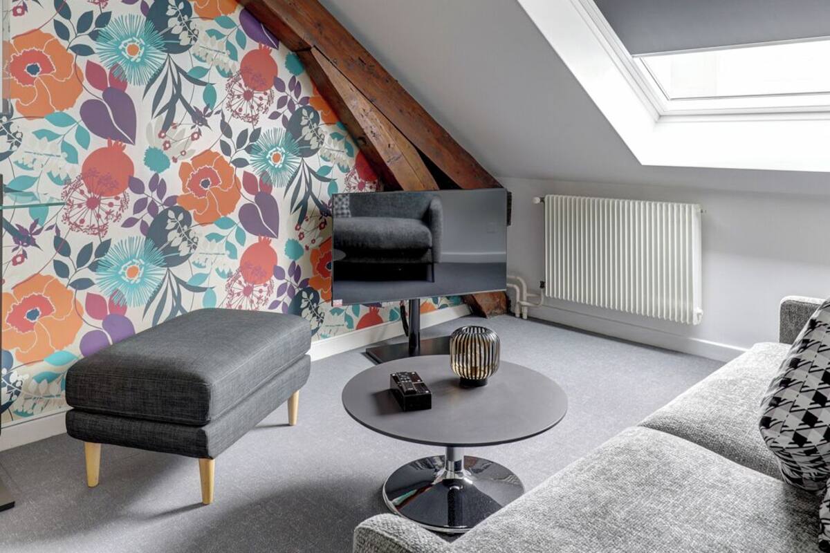 Een moderne loft slaapkamer met een schuine plafond, dakraam, kleurrijke bloemen behang, een grijze bank, een ronde salontafel en een grijze fauteuil.