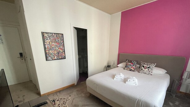 Habitación de hotel con cama, control remoto, puerta y pared con un cuadro de colores.