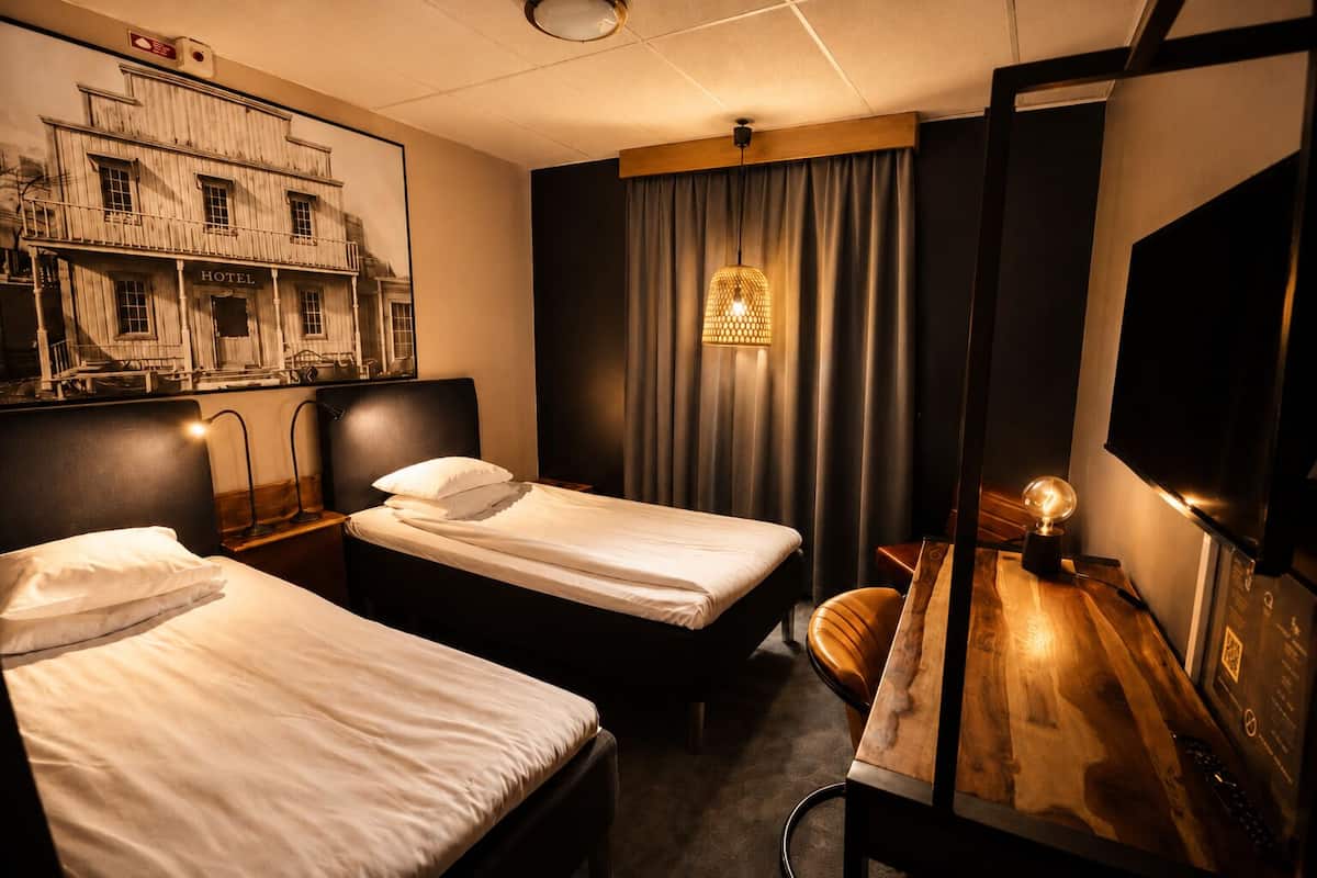 A hotel room with two beds, a desk, a chair, a TV, and a wall-mounted picture of a building.