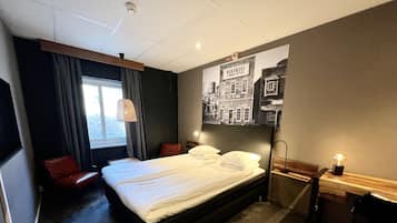 A hotel room with a large bed, a red chair, a desk with a lamp, and a window with curtains.