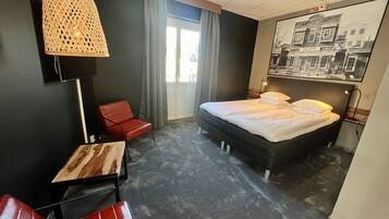 Une chambre d’hôtel moderne avec un grand lit, un fauteuil rouge, une petite table, un luminaire suspendu et un tableau représentant un paysage urbain accroché au mur.