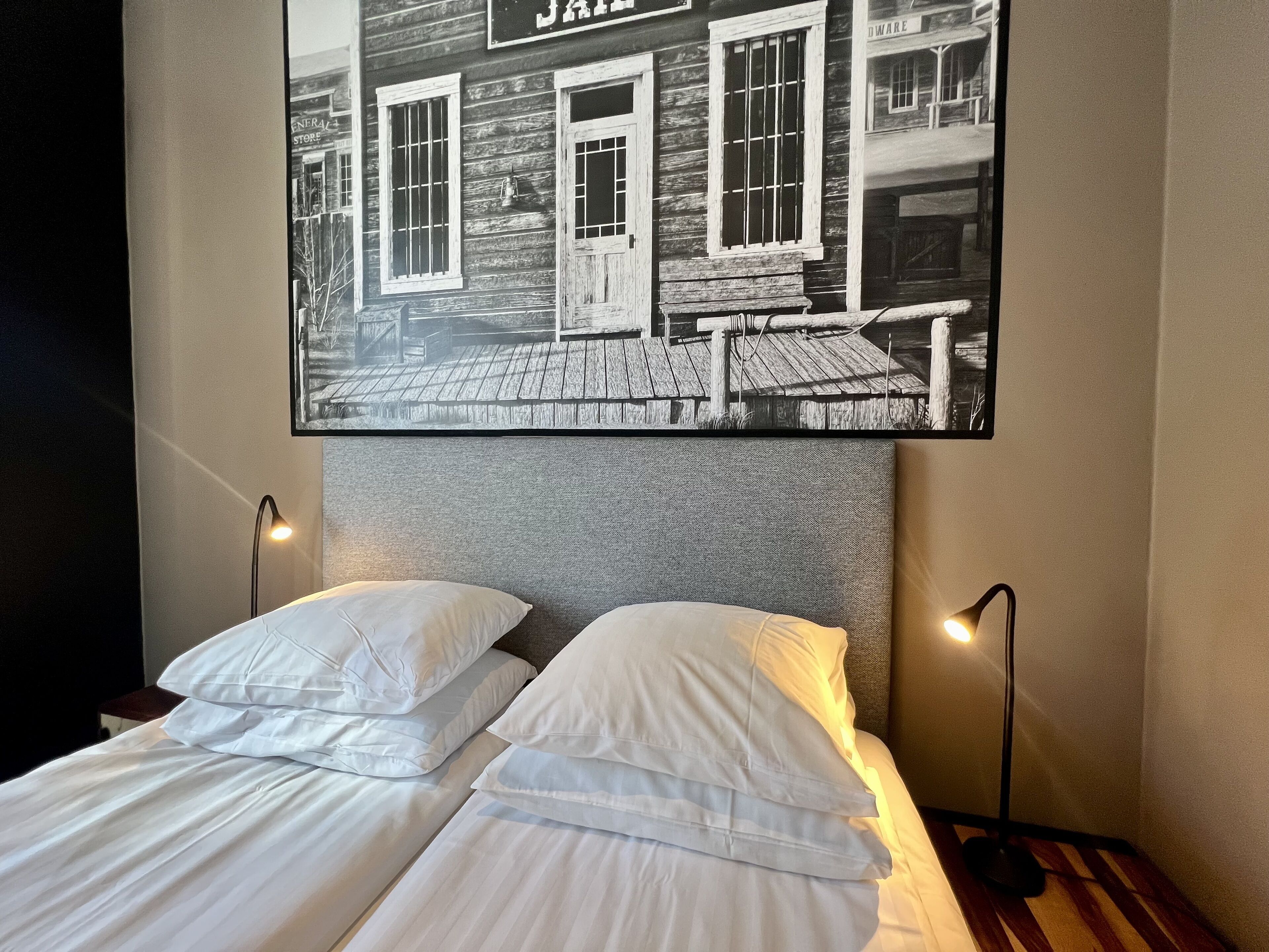 A hotel room with two beds, each with white linens and pillows, and a large black and white photo of a building with barred windows and a sign that reads "JAMES".