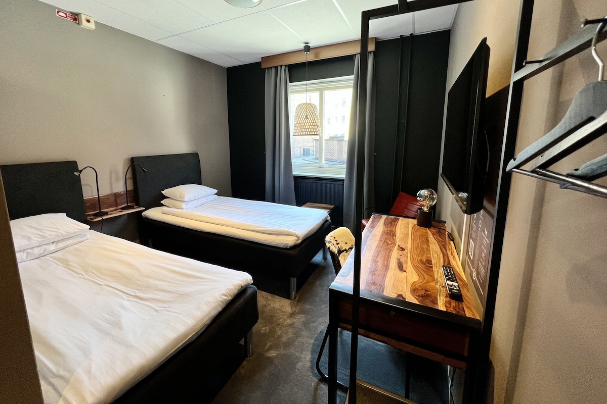 Una habitación de hotel con dos camas, un escritorio de madera, un televisor montado en la pared y una ventana con cortinas.