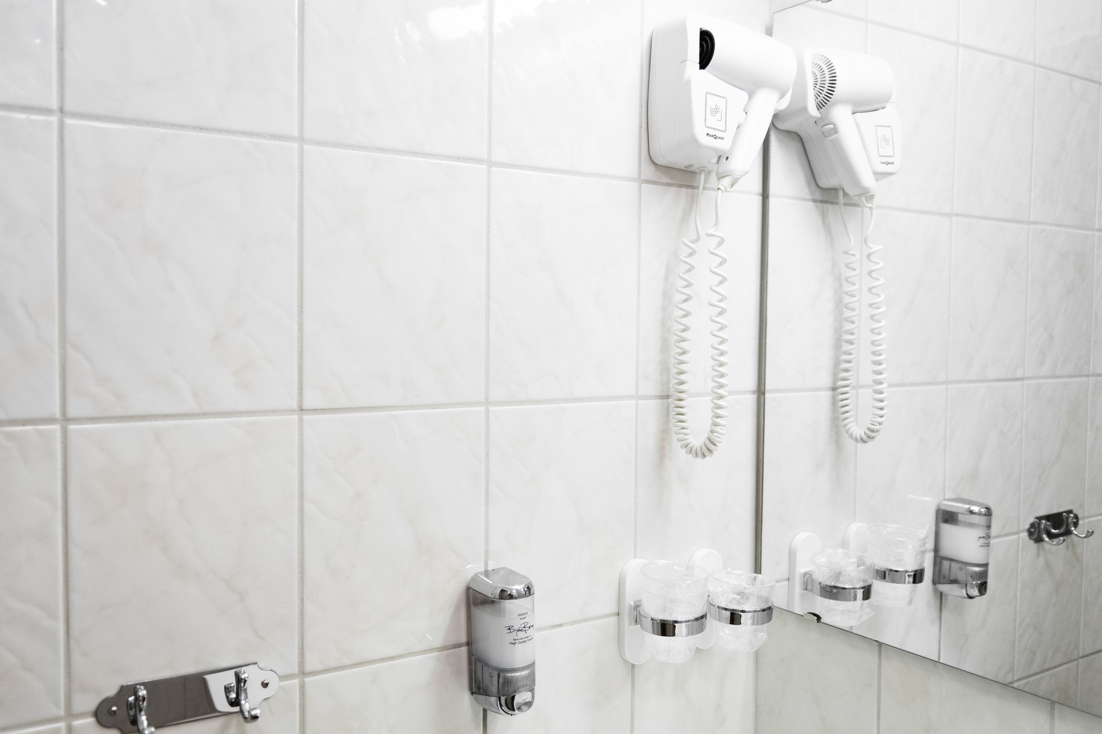 A bathroom with white tiles, a hairdryer, a mirror, and a soap dispenser.