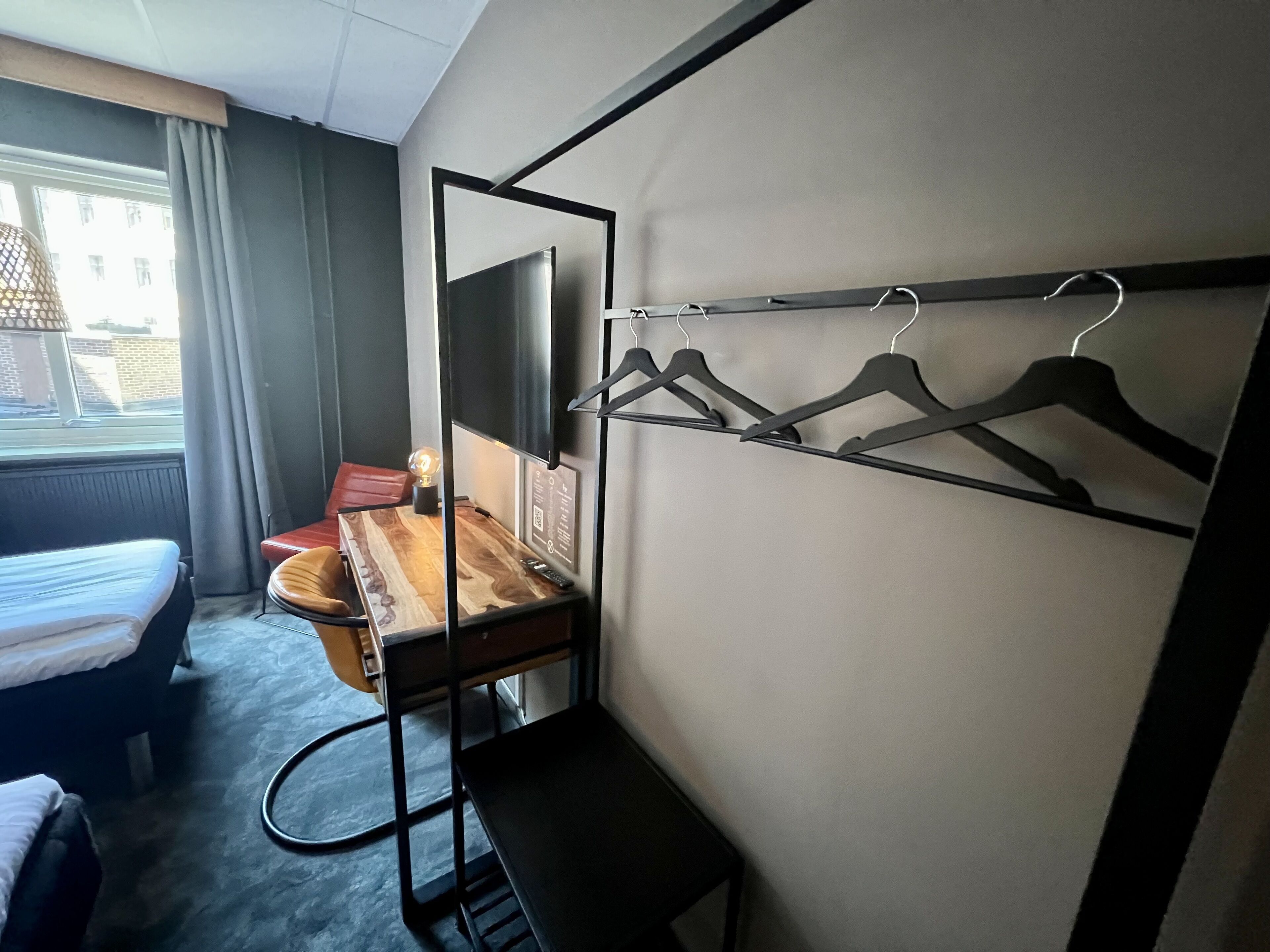 A hotel room with a bed, a desk with a mirror, a chair, a coat rack, and a view of buildings outside the window.