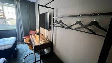 A hotel room with a bed, a desk with a mirror, a chair, a coat rack, and a view of buildings outside the window.