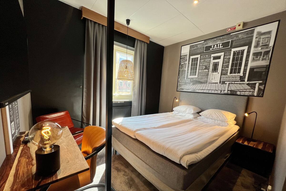 A hotel room with a bed, a desk with a lamp, a chair, and a wall-mounted picture.