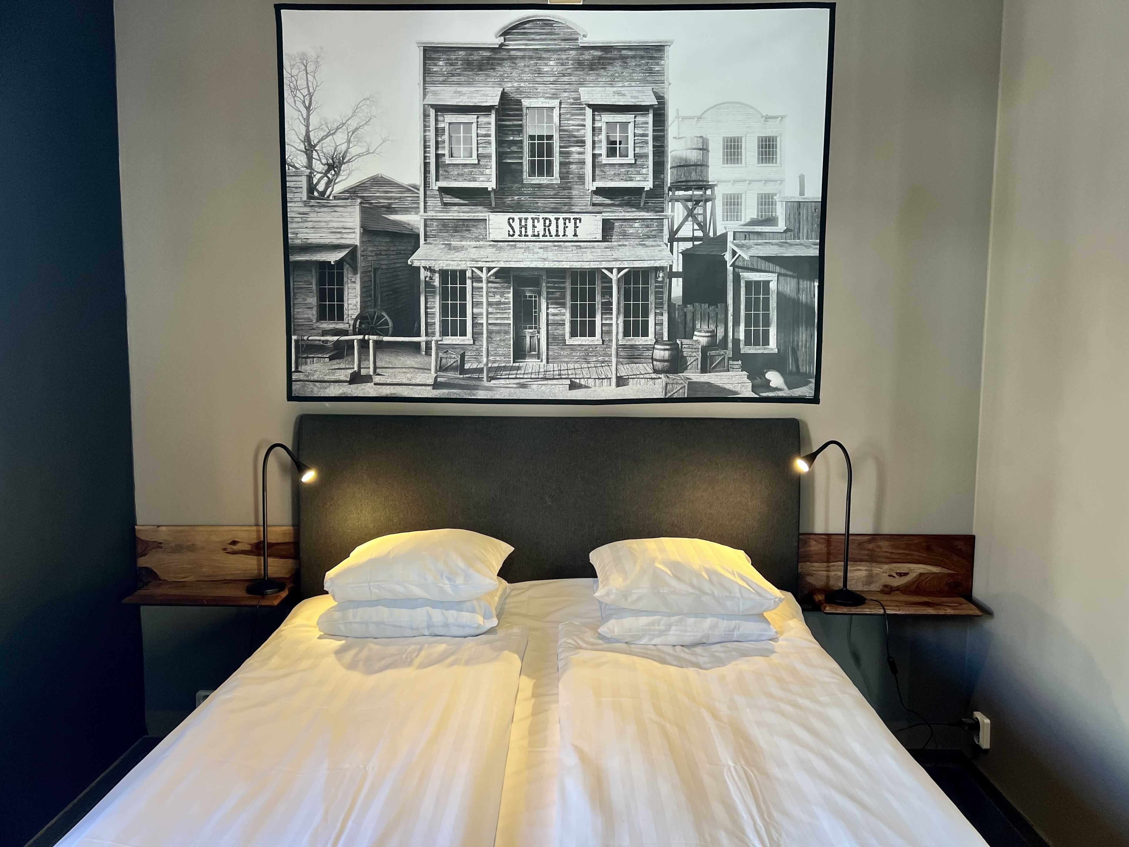 A bed with white linens and two pillows, a headboard with two wall-mounted lamps, and a framed black and white photograph of a building labeled "SHERIFF" above the bed.