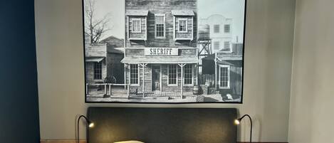 A bed with white linens and two pillows, a headboard with two wall-mounted lamps, and a framed black and white photograph of a building labeled "SHERIFF" above the bed.