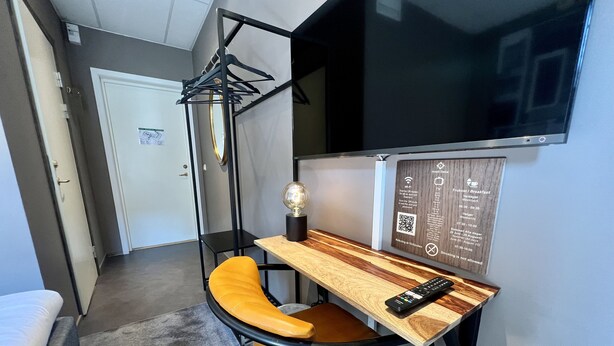 Habitación de hotel compacta con una cama individual, un escritorio pequeño con una silla, un televisor montado en la pared e información sobre servicios en un panel de madera.