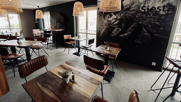 A modern restaurant interior with wooden tables and chairs, hanging wicker lamps, and a large mural on the wall. - Hotell Siesta (Karlskrona)