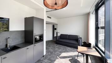 Cozinha moderna com micro-ondas embutido, pia e luminária pendente. Na sala de estar, um sofá escuro e uma mesa pequena com cadeiras, além de uma grande janela que proporciona luz natural.