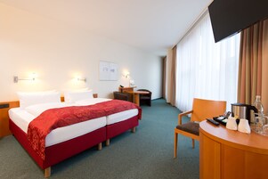 Una habitación de hotel con cama, escritorio, silla, televisión y ventana con cortinas.