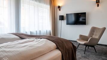 A hotel room with a large bed, a chair, a flat-screen TV, and a window with sheer curtains.
