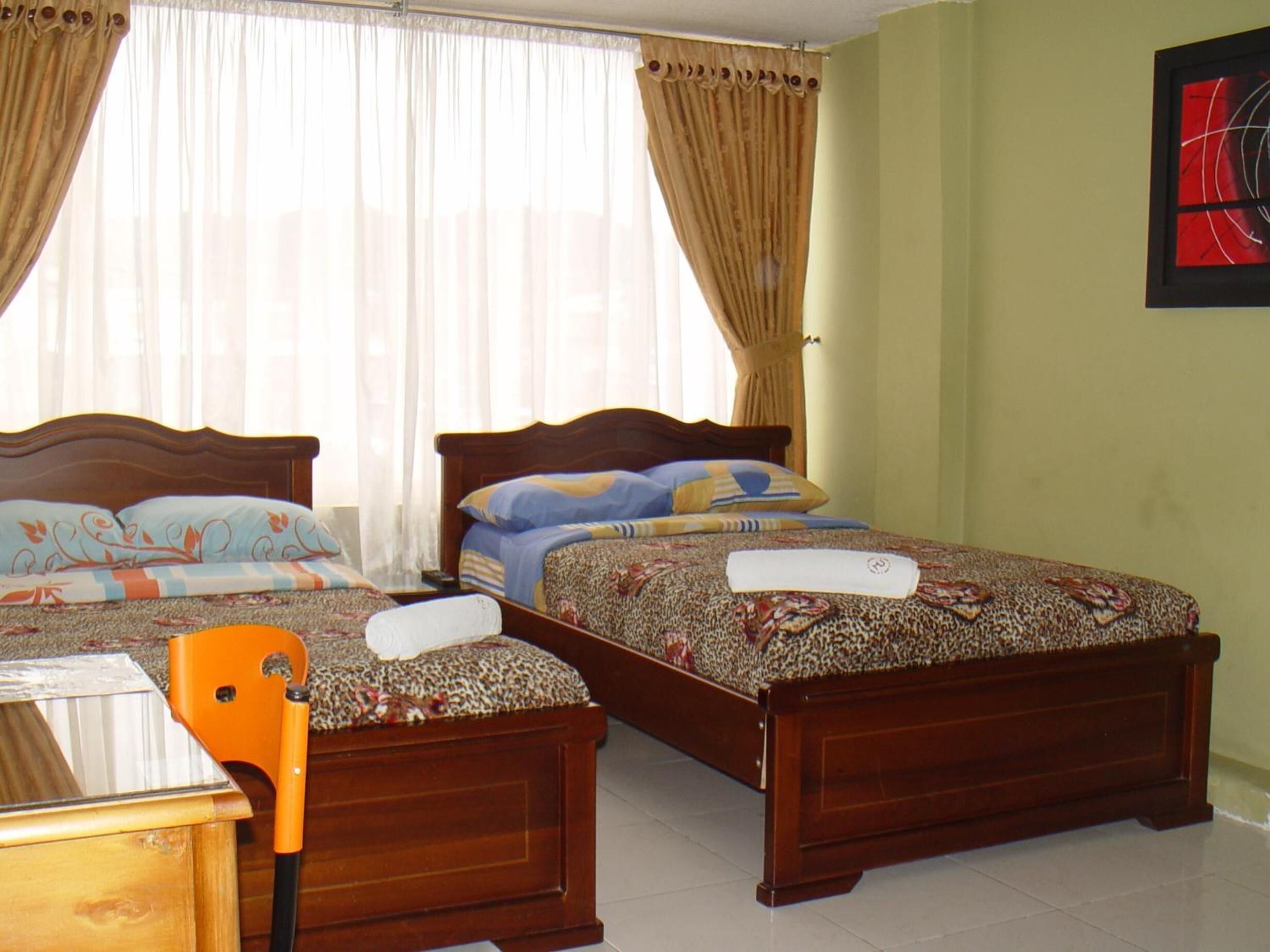 Habitación de hotel con dos camas de madera, una mesita de noche de madera, una silla y una ventana con cortinas translúcidas.