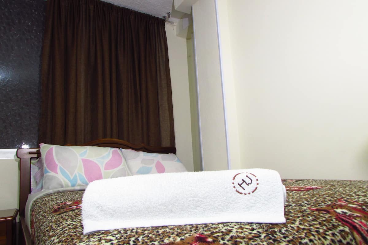 A neatly made bed with a patterned bedspread, a pillow with a colorful design, and a brown curtain partially drawn.