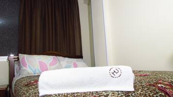 A neatly made bed with a patterned bedspread, a pillow with a colorful design, and a brown curtain partially drawn.